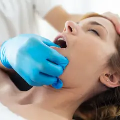 Fizjoterapeuta w niebieskiej rękawiczce delikatnie bada szczękę pacjentki, która cierpi na **szczekoscisk**.