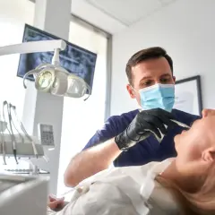 Dentysta w masce wykonuje zabieg szynowania zębów u pacjentki.
