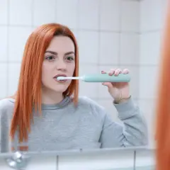 Kobieta z rudymi włosami myje zęby zieloną szczoteczką soniczną przed lustrem.