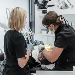 Dwie dentystki pracują przy pacjencie. Jedna używa mikroskopu, druga trzyma narzędzie. To obrazek o tym, jak zostać dentystą.