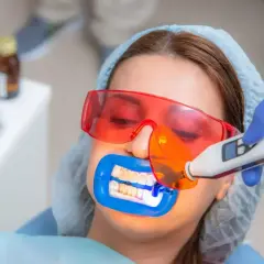 Zabieg lakowania zębów uśmiechniętej pacjentki w okularach ochronnych. Dentysta utwardza lak światłem lampy.