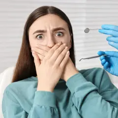 Przerażona pacjentka z dentofobią zakrywa usta. W pobliżu widać ręce dentysty w niebieskich rękawiczkach z narzędziami.