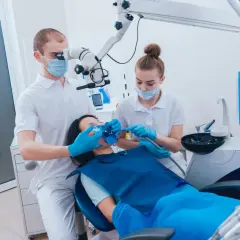 Dentysta pod mikroskopem bada ząb pacjentki, która leży na fotelu. Asystentka przygotowuje się do zabiegu, by zapobiec zatruciu zęba.