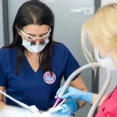 Dwie dentystki w maseczkach i okularach pracują przy pacjencie. Jedna używa wiertła, druga odsysacza. To profesjonalna artykaina.