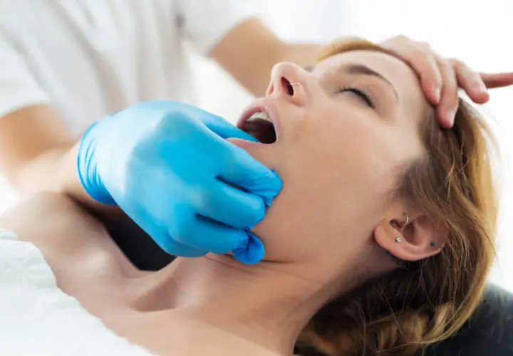 Fizjoterapeuta w niebieskiej rękawiczce delikatnie bada szczękę pacjentki, która cierpi na **szczekoscisk**.