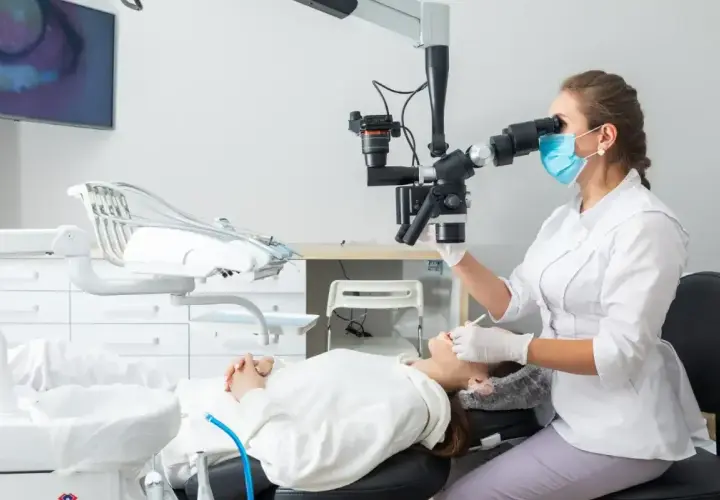 Dentystka w masce i rękawiczkach wykonuje zabieg sanacji przy użyciu mikroskopu. Pacjentka leży na fotelu dentystycznym.