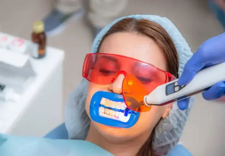 Zabieg lakowania zębów uśmiechniętej pacjentki w okularach ochronnych. Dentysta utwardza lak światłem lampy.