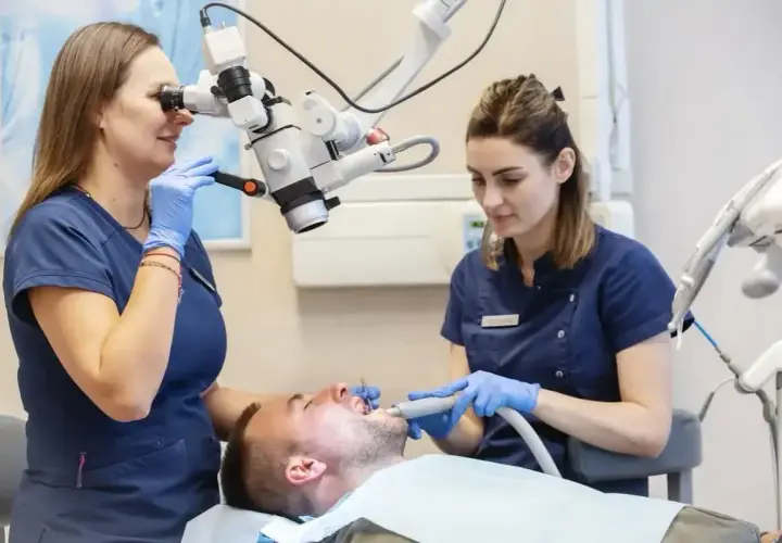 Dwie dentystki w gabinecie stomatologicznym pracują przy pacjencie, jedna używa mikroskopu, druga narzędzia.