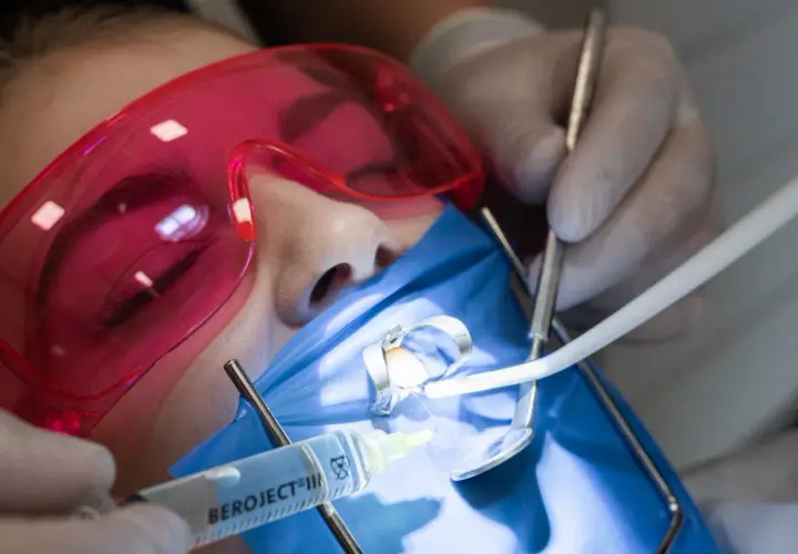 Precyzyjne leczenie kanałowe pod mikroskopem. Dentysta w rękawiczkach używa narzędzi i strzykawki. Pacjent ma założone różowe okulary ochronne.