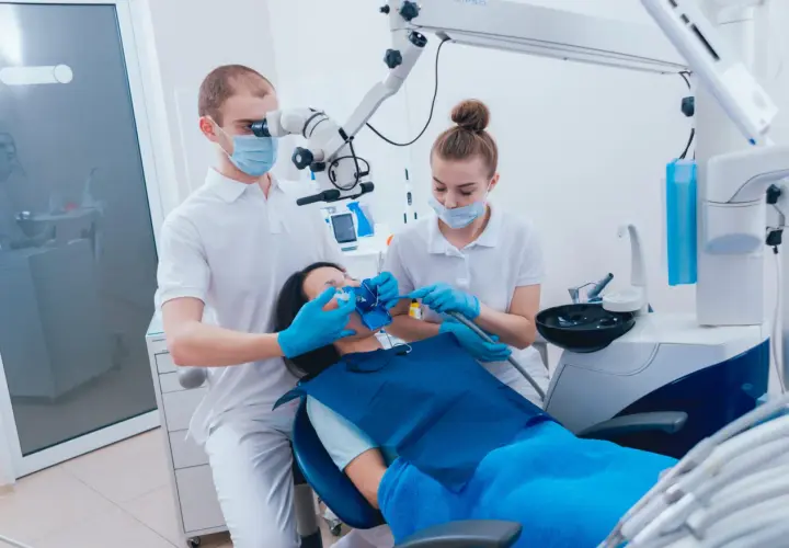 Dentysta pod mikroskopem bada ząb pacjentki, która leży na fotelu. Asystentka przygotowuje się do zabiegu, by zapobiec zatruciu zęba.