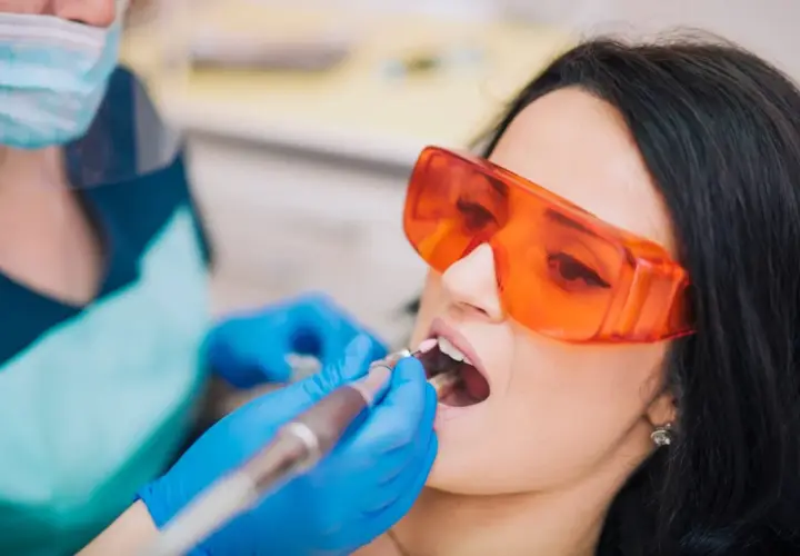 Pacjentka w okularach ochronnych poddaje się zabiegowi stomatologicznemu. Dentysta w rękawiczkach używa wiertła. Nowoczesne znieczulenie komputerowe zapewnia komfort.
