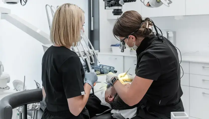 Dwie dentystki pracują przy pacjencie. Jedna używa mikroskopu, druga trzyma narzędzie. To obrazek o tym, jak zostać dentystą.