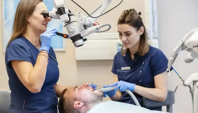 Dwie dentystki w gabinecie stomatologicznym pracują przy pacjencie, jedna używa mikroskopu, druga narzędzia.