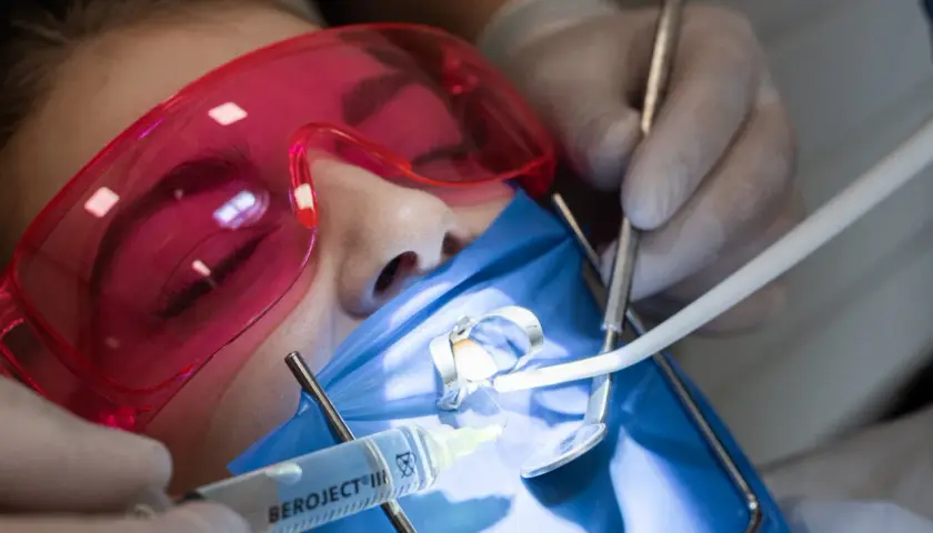 Precyzyjne leczenie kanałowe pod mikroskopem. Dentysta w rękawiczkach używa narzędzi i strzykawki. Pacjent ma założone różowe okulary ochronne.