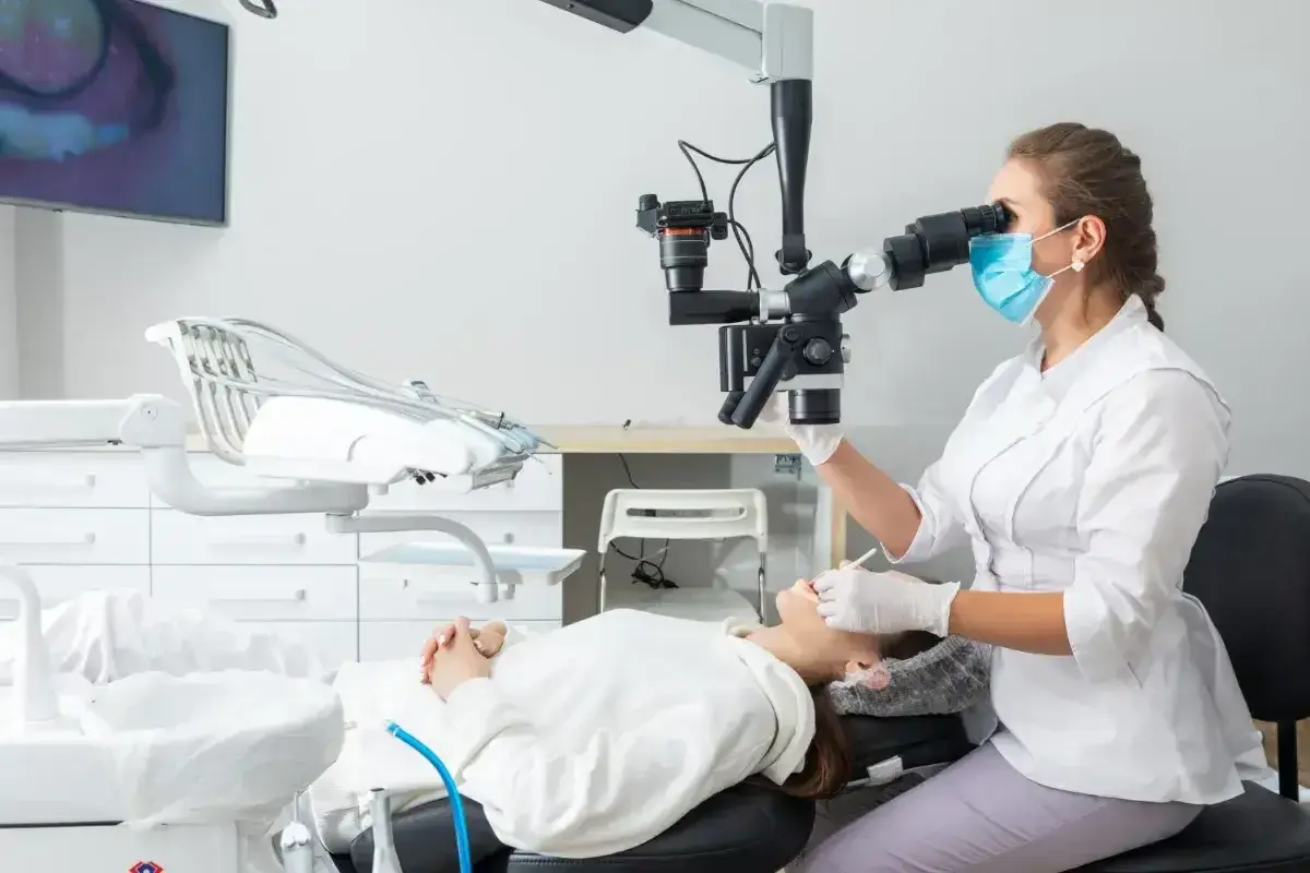 Dentystka w masce i rękawiczkach wykonuje zabieg sanacji przy użyciu mikroskopu. Pacjentka leży na fotelu dentystycznym.