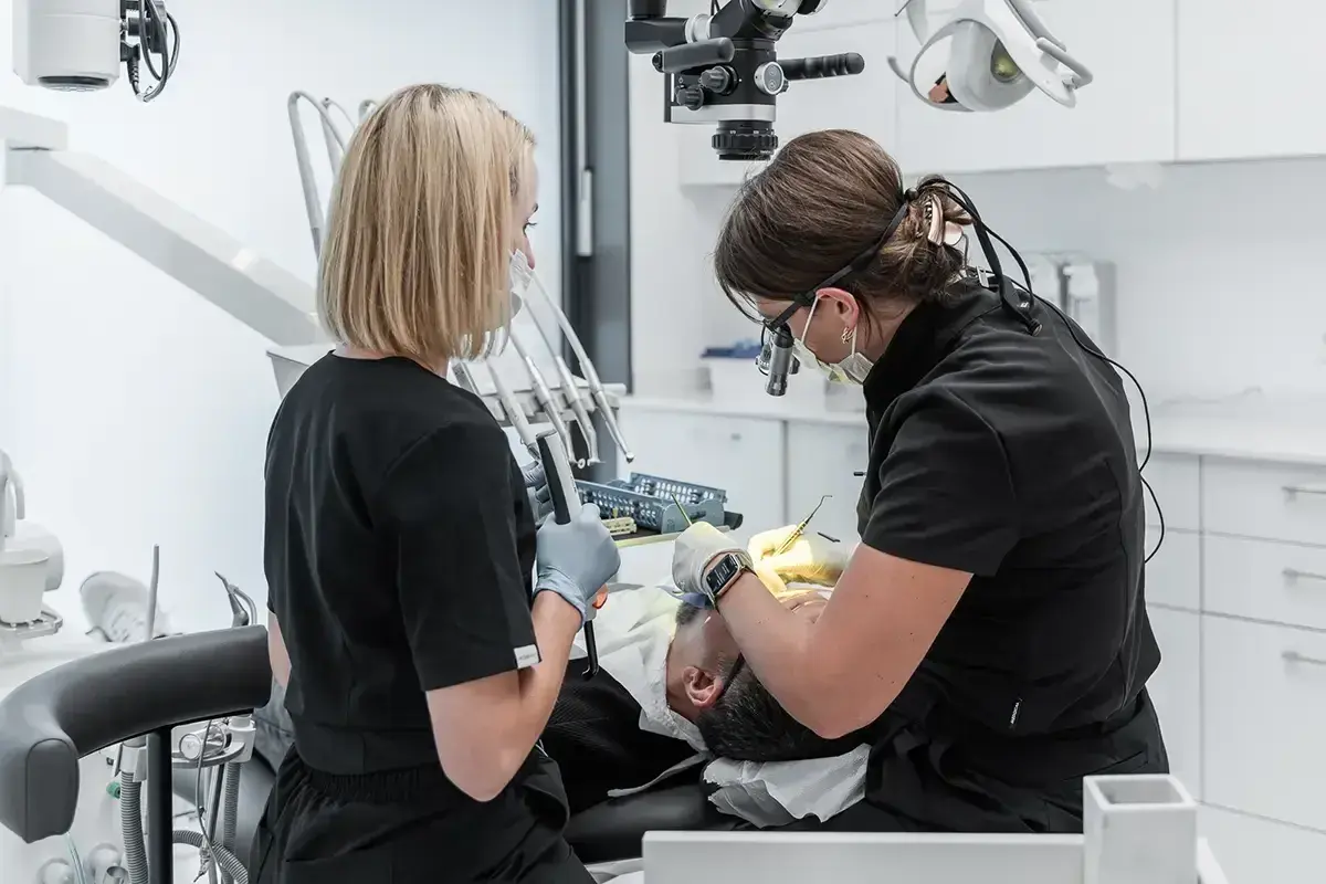 Dwie dentystki pracują przy pacjencie. Jedna używa mikroskopu, druga trzyma narzędzie. To obrazek o tym, jak zostać dentystą.