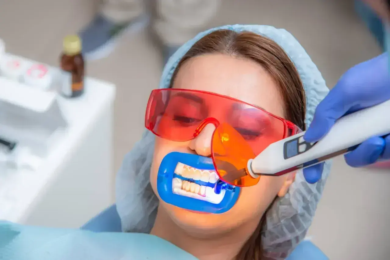 Zabieg lakowania zębów uśmiechniętej pacjentki w okularach ochronnych. Dentysta utwardza lak światłem lampy.