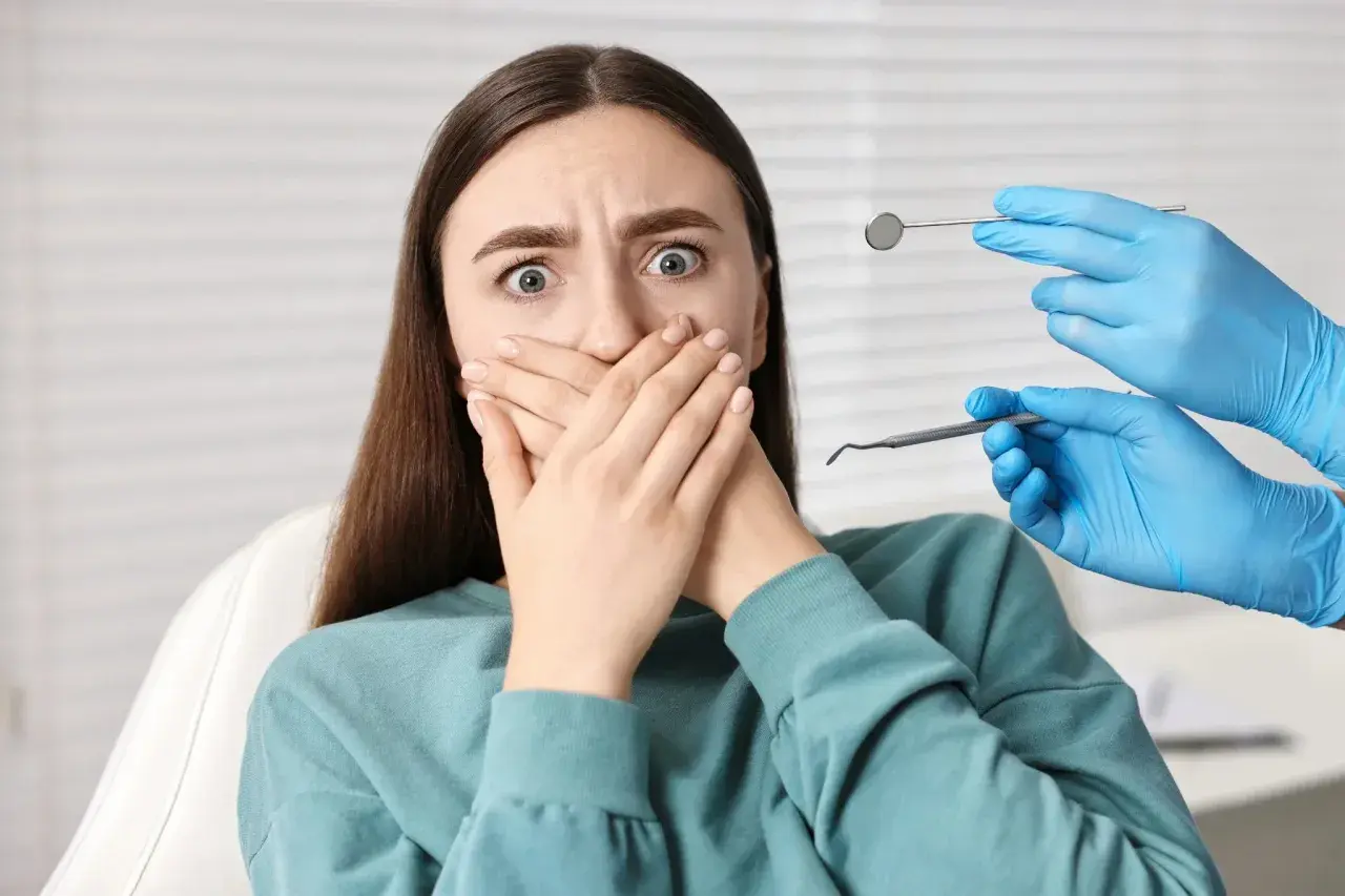 Przerażona pacjentka z dentofobią zakrywa usta. W pobliżu widać ręce dentysty w niebieskich rękawiczkach z narzędziami.
