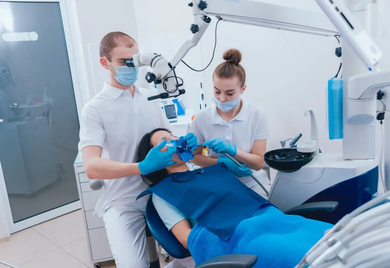 Dentysta pod mikroskopem bada ząb pacjentki, która leży na fotelu. Asystentka przygotowuje się do zabiegu, by zapobiec zatruciu zęba.