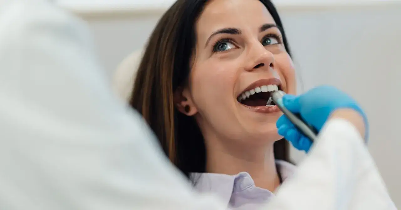 Uśmiechnięta kobieta podczas zabiegu borowania zębów. Dentysta w niebieskich rękawiczkach używa wiertła.