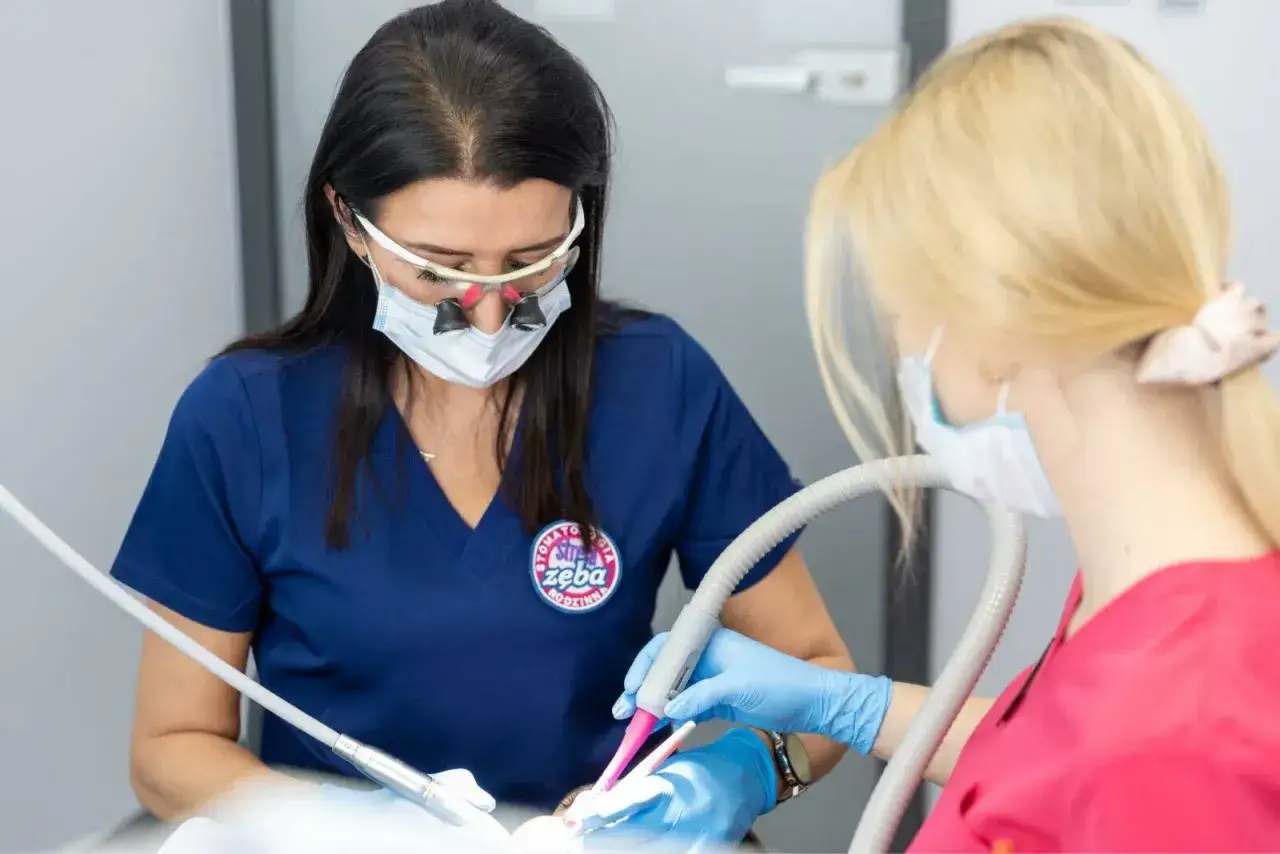 Dentystka w okularach powiększających i masce wykonuje zabieg. Warto znać lidokaina skutki uboczne.