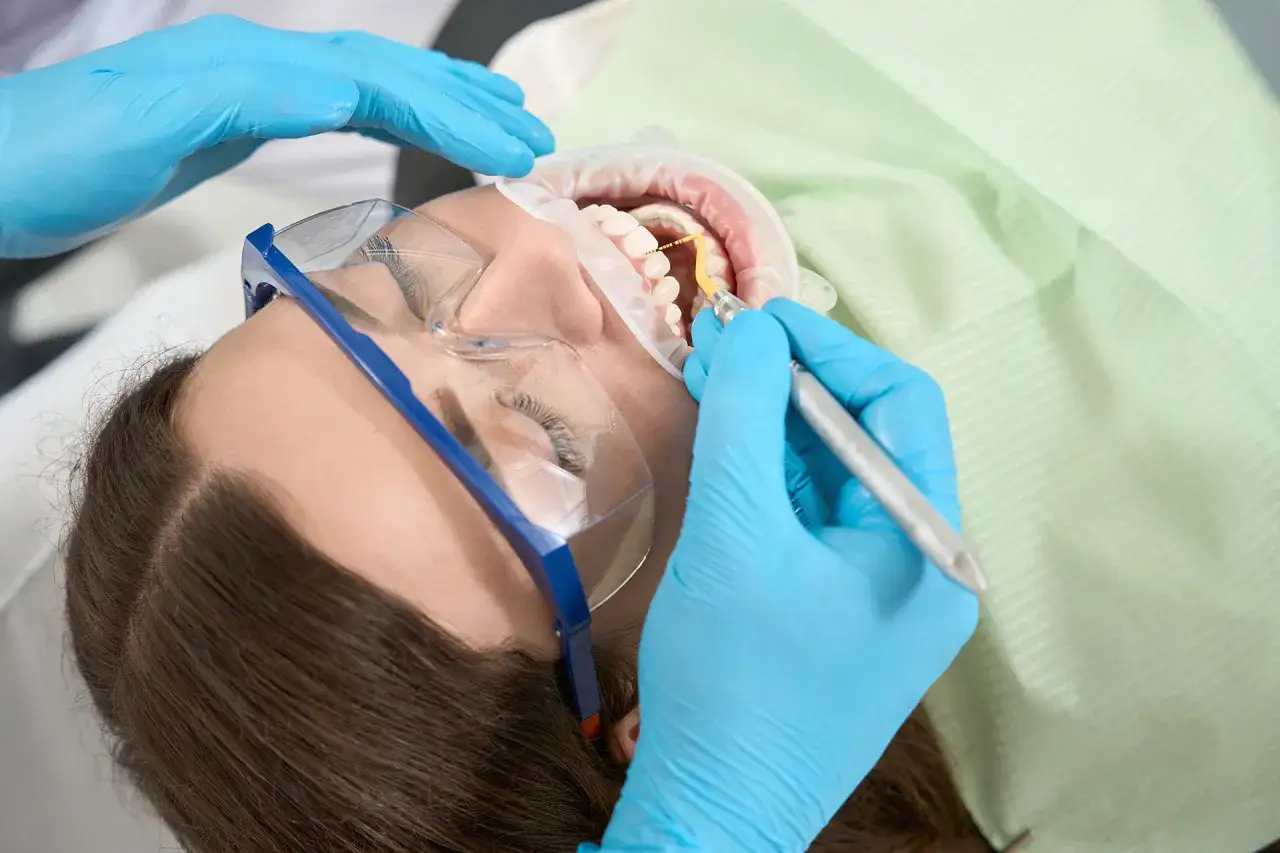 Profesjonalna higienizacja zębów. Dentysta w niebieskich rękawiczkach używa narzędzia do czyszczenia uzębienia pacjentki.