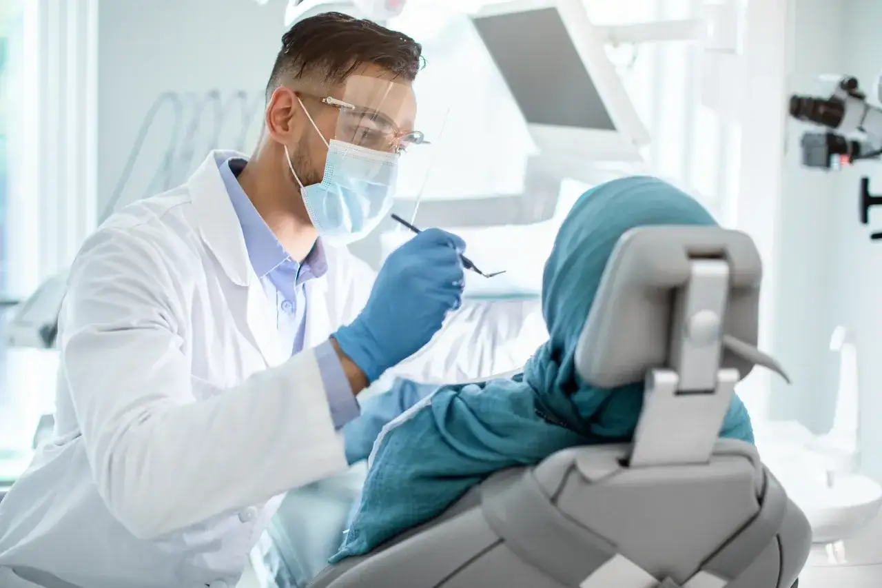 Dentysta w masce i fartuchu bada pacjentkę. Zastanawiasz się, ile zarabia dentysta NFZ?