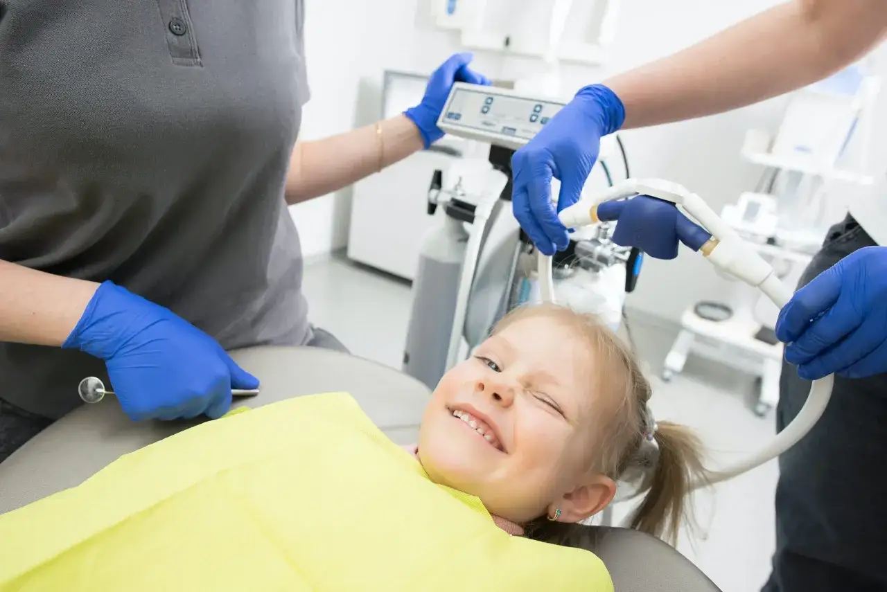 Uśmiechnięta dziewczynka na fotelu dentystycznym, podczas gdy dentysta przygotowuje sprzęt. W tle widać urządzenie z panelem sterowania, które może służyć do podawania gazu rozweselającego (mih).