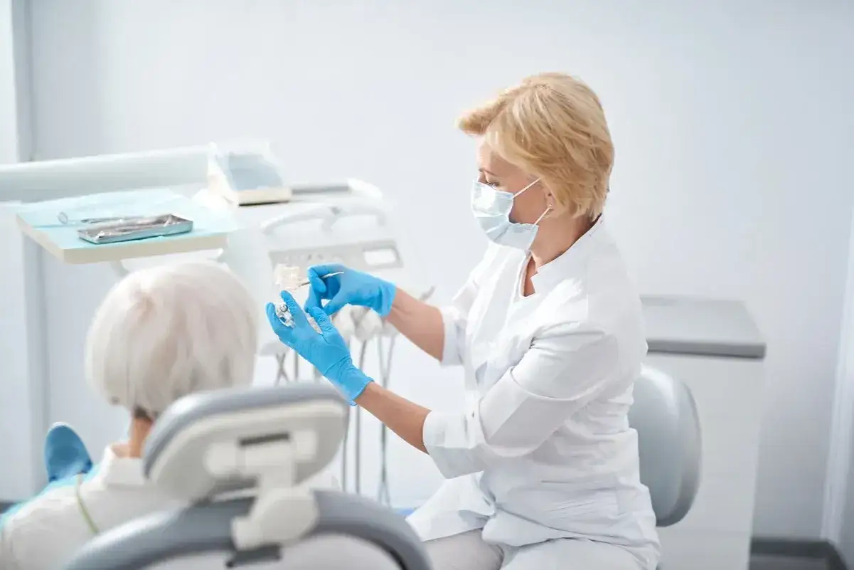Dentystka w maseczce i rękawiczkach pokazuje pacjentowi model szczęki. Mediraty opinie potwierdzają profesjonalizm.